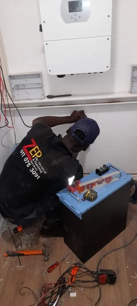 Technician in a ZEPS shirt kneeling beside a battery pack under a wall-mounted inverter, with tools on the floor