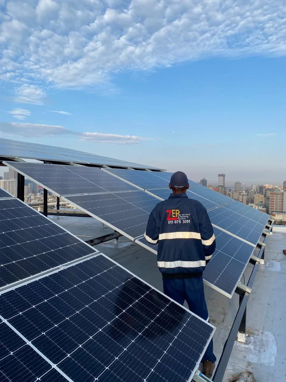 Technician in a ZEPS navy jacket standing among rooftop solar panels, looking out over the Johannesburg skyline