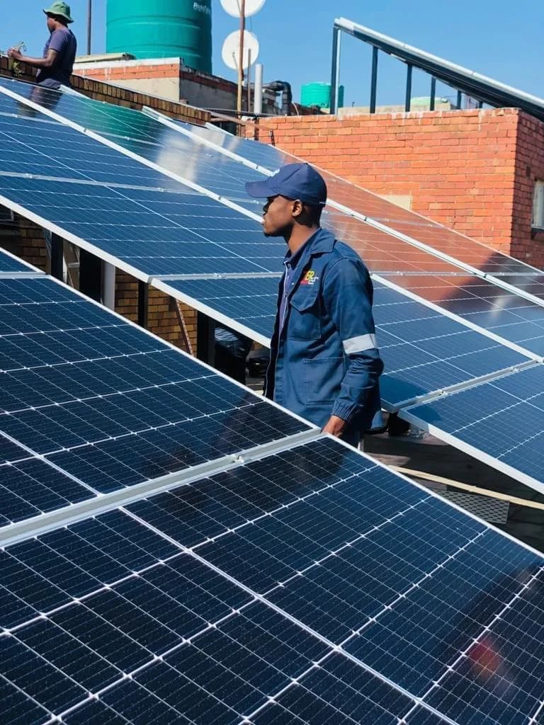 Technician in a navy cap and jacket standing among rooftop solar panels, with another crew member by a brick wall